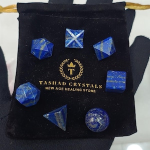 May include: Set of seven blue lapis lazuli gemstone dice in a velvet pouch. The dice are in the shapes of a sphere, pyramid, cube, and a few other geometric shapes. The pouch is black with gold lettering that reads "Tashadd Crystals New Age Healing Stone".
