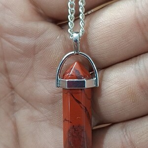 May include: A silver chain necklace with a red jasper crystal pendant. The pendant is a point shape and is set in a silver bezel.