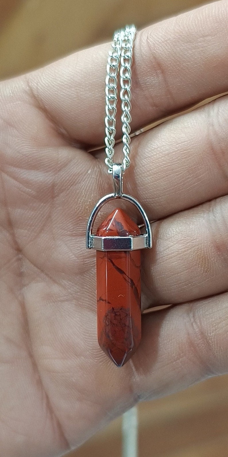 May include: A silver chain necklace with a red jasper crystal pendant. The pendant is a point shape and is set in a silver bezel.