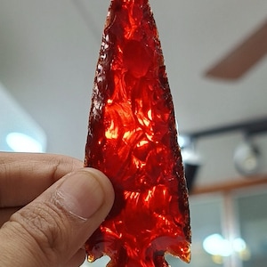 May include: A red obsidian arrowhead, a type of volcanic glass. The arrowhead is translucent and has a sharp point.