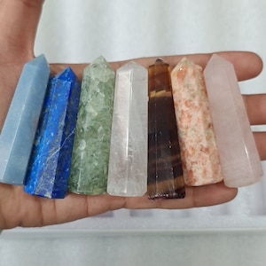 Op de afbeelding: Zeven verschillende gekleurde kristallen staven, waaronder blauw, groen, wit, bruin, oranje en roze. De staven zijn in een rij gerangschikt en worden in een hand gehouden.