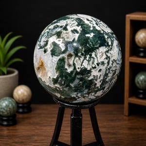May include: A large, polished moss agate sphere, predominantly white with green and brown inclusions, sits on a black metal stand. The sphere is displayed on a wooden surface, with other decorative spheres in the background. The agate sphere is a decorative item.