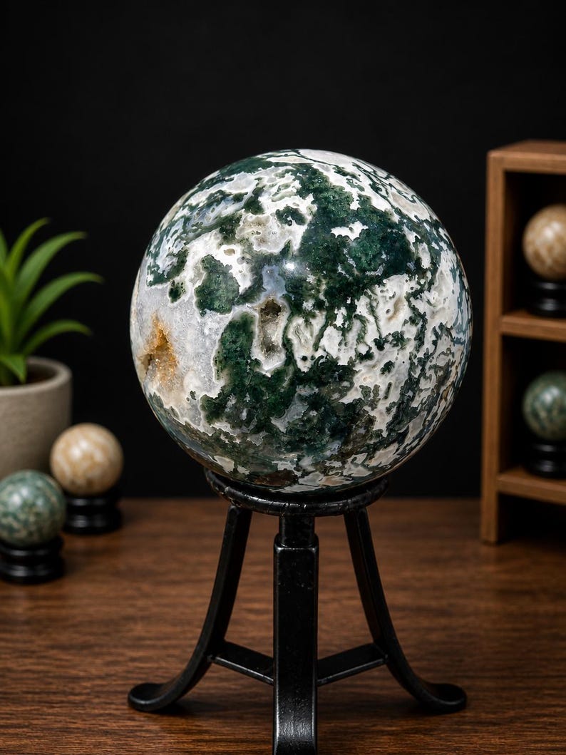 May include: A large, polished moss agate sphere, predominantly white with green and brown inclusions, sits on a black metal stand. The sphere is displayed on a wooden surface, with other decorative spheres in the background. The agate sphere is a decorative item.