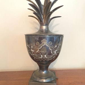 May include: A metallic pineapple-shaped container with a removable lid featuring pointed leaves. The body is dark with engraved floral patterns and sits on a square base with four small feet. The object has a polished finish with visible patina.