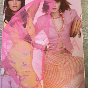 May include: A rectangular Escada Spring/Summer 2003 fashion catalog featuring two models. One wears a yellow floral dress with a pink handbag, while the other wears a pink textured jacket over a yellow and pink gingham skirt with a tan handbag.
