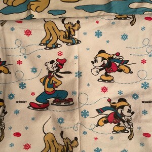 May include: White fabric featuring cartoon characters Mickey Mouse and Pluto ice skating. The design includes red and blue snowflakes, hockey pucks, and dotted lines. The Disney logo is visible.