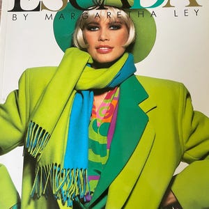 May include: A vintage fashion advertisement featuring the text ESCADA BY MARGARETHA LEY. The image displays a lime green blazer, a matching green hat, and a two-toned scarf in bright blue and lime green, layered over a colorful patterned shirt.