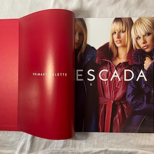 May include: A red and white book with the word "EScada" printed in white on the cover. The book is open to a page with a photo of three women wearing winter coats.