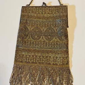 May include: A rectangular beaded handbag featuring a metallic gold and bronze geometric pattern. The bag has a metal frame with a kiss-lock closure, a thin chain handle, and a long fringe of matching metallic beads along the bottom edge.