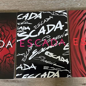 May include: Three rectangular booklets with the word ESCADA printed on each. The left booklet features a red and black rose pattern. The center booklet has a black background with white repeating ESCADA text. The right booklet has a red zebra print.