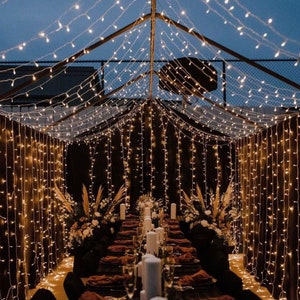 May include: A long dining table set with white candles and brown placemats is decorated with white flowers and greenery. The table is set under a canopy of fairy lights, creating a warm and inviting atmosphere.