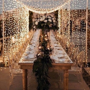 May include: A long wooden table with a white tablecloth is set for a wedding reception. The table is decorated with a lush green garland and white flowers. The table is surrounded by clear plastic chairs and a string of fairy lights hangs above the table.