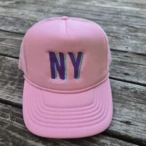Puede incluir: Gorra trucker rosa con parte trasera de malla y visera curva. La parte delantera de la gorra presenta las letras "NY" en morado con una sombra azul claro. La gorra está hecha de un material suave.