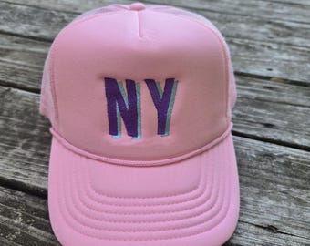May include: A pink trucker hat with a mesh back and a curved brim. The front of the hat features the letters "NY" in purple with a light blue shadow. The hat is made of a soft material and has a classic trucker hat design.