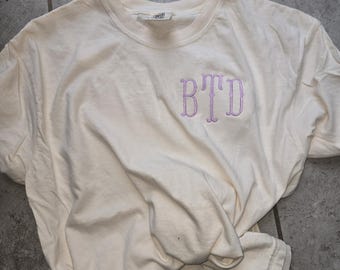 Puede incluir: Una camiseta blanca con un monograma bordado en morado que dice "BTD".