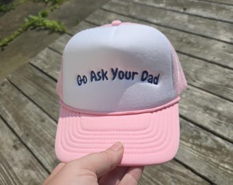 Könnte beinhalten: Rosa-weiße Trucker-Cap mit dem Text "Go Ask Your Dad" in Marineblau gestickt. Die Cap hat einen rosa Schirm und ein Mesh-Rückenteil sowie eine weiße Frontplatte.