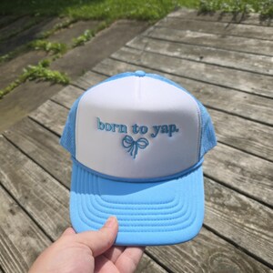 May include: A light blue and white trucker hat with the embroidered text "born to yap." The hat features a curved brim and a mesh back. The text is in a light blue color, and there is a bow graphic below the text.