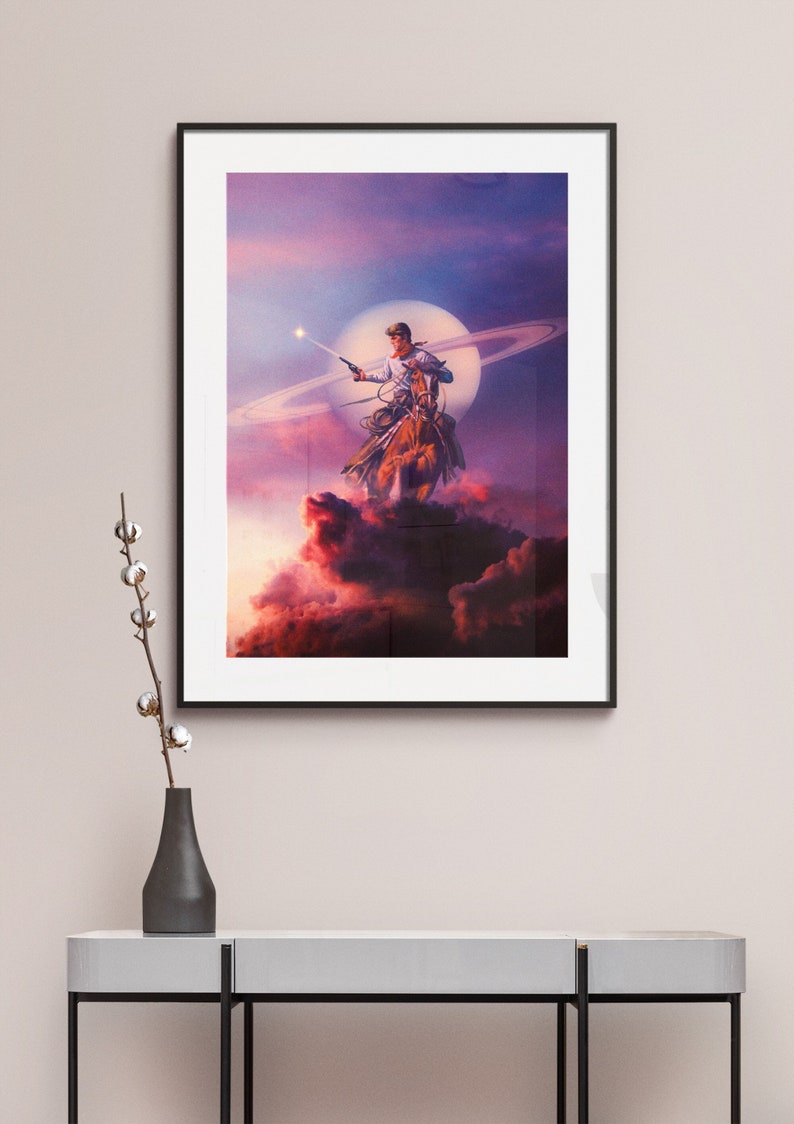 May include: A framed poster featuring a cowboy riding a horse in front of a large, bright moon and a ring of planets. The cowboy is holding a gun and pointing it towards the sky. The background is a vibrant purple and pink.
