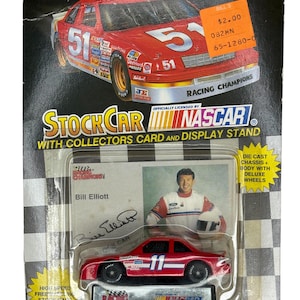 Puede incluir: Un coche de carreras NASCAR rojo con el número 11, empaquetado con una tarjeta de coleccionista con Bill Elliott. El embalaje incluye los logotipos de Racing Champions y NASCAR, y el texto "Die Cast Chassis + Body with Deluxe Wheels."