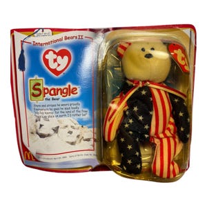 May include: A collectible Ty Beanie Baby bear named Spangle, packaged in a clear plastic case. The bear's body is decorated with a patriotic design of red and white stripes and blue stars. The packaging includes the text "International Bears II" and a poem.