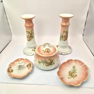 May include: A collection of vintage porcelain items, including two candle holders, a lidded jar, and two decorative dishes. The pieces feature a peach and white color scheme with floral designs and gold trim, creating an elegant aesthetic.