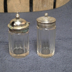 Puede incluir: Dos frascos de vidrio vintage con tapas de metal plateado. Una tapa tiene una pequeña abertura para cuchara, la otra tiene pequeños agujeros. Los frascos son transparentes con estrías verticales. Los frascos son probablemente para especias o condimentos.