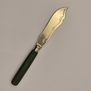May include: A butter knife with a curved, gold-colored blade featuring an etched floral design. The handle is dark green and has a silver-colored band where it meets the blade. The knife is designed for spreading butter or soft cheeses.