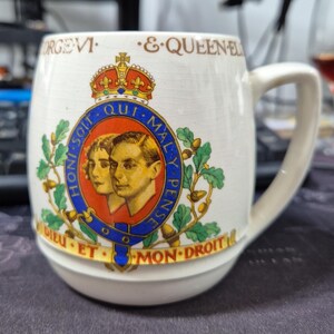 Może przedstawiać: Biały ceramiczny kubek z królewskim herbem z portretami Jerzego VI i królowej Elżbiety. Herb zawiera koronę, liście dębu i motto "Dieu et mon droit". Kubek ma zakrzywiony uchwyt i lekko rozszerzoną podstawę.