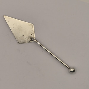 May include: A silver-colored cake server with a diamond-shaped blade and a long, thin handle. The handle ends in a small, round ball. The server is designed for serving cakes and pastries.