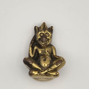 Puede incluir: Pequeña figura de latón de un duende sentado con una corona. El duende tiene una cara sonriente y está en posición de piernas cruzadas. La figura tiene un aspecto envejecido y está sobre un fondo blanco.