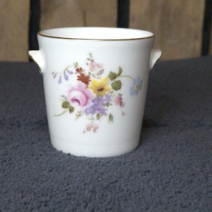 Puede incluir: Jarrón de cerámica blanca con un diseño floral con flores rosas, amarillas y moradas. El jarrón tiene un borde dorado y asas pequeñas. Una pieza decorativa para el hogar.