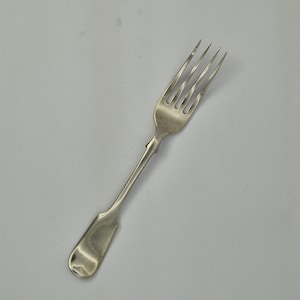 May include: A silver fork with four tines and a decorative handle. The fork has a rectangular base and a slender, curved handle. The tines are evenly spaced and slightly curved. The fork is made of a shiny, reflective metal.