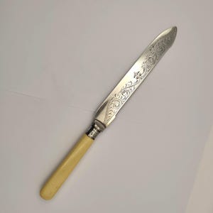 May include: A silver butter knife with an ivory-colored handle. The blade is engraved with a floral pattern. The knife is approximately 8 inches long.