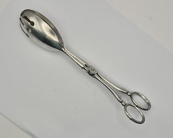 May include: Silver-toned serving tongs with a slotted oval head and scissor-style handles. The handles have decorative detailing and end in two finger loops. The tongs are designed for serving food.