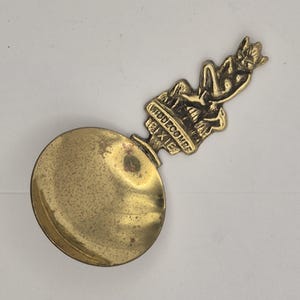 May include: A vintage brass spoon with a round bowl and a handle featuring a reclining pixie figure. The handle also has the text "WIDDECOMBE PIXIE". The spoon has a tarnished, aged appearance.