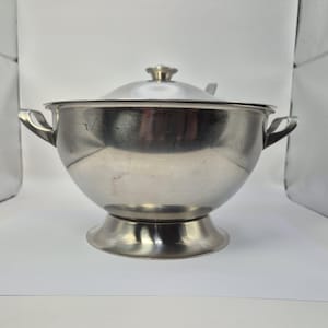 Könnte beinhalten: Eine Edelstahlsuppenterrine mit Deckel und zwei Griffen. Die Terrine hat einen Sockel und ein Löffel liegt auf dem Deckel. Das Metall hat eine gebürstete Oberfläche und reflektiert das Umgebungslicht.