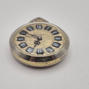 May include: A silver and gold Trafalgar pocket watch with a gold-tone face. The watch features black Roman numerals and hands, with the word "Trafalgar" printed on the face. A classic timepiece.