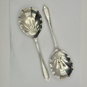 Puede incluir: Dos cucharas de servir de plata antiguas. Los cuencos de las cucharas tienen un diseño de concha con líneas radiales. Los mangos son largos y delgados, con un borde decorativo. Las cucharas se muestran sobre un fondo blanco.