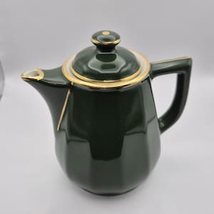 May include: A dark green ceramic teapot with a gold rim and handle. The teapot has a matching lid with a gold accent. The spout is angled and the handle is square. The teapot is on a white background.