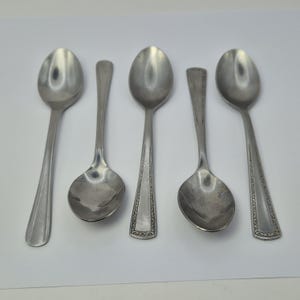 May include: Five vintage silver spoons are displayed on a white surface. The spoons have different designs, with oval bowls and long handles. Some handles feature decorative patterns. The spoons are made of a shiny, reflective metal.