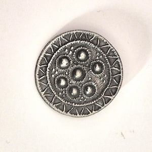 May include: A detailed, silver-toned metal button with an intricate design. The button features a central cluster of raised circles surrounded by concentric rings of textured patterns and triangular shapes. The button is circular and has a slightly aged appearance.