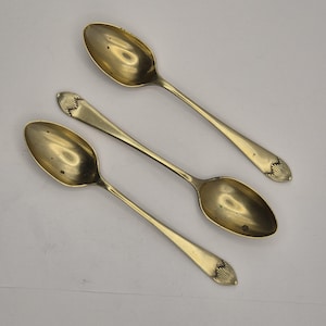 Pode incluir: Três colheres antigas douradas dispostas diagonalmente sobre uma superfície branca. Cada colher tem uma tigela oval e um cabo longo e fino. A alça termina em um design decorativo em forma de leque. As colheres parecem ser de metal.