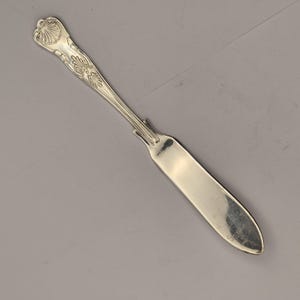 Può includere: Coltello da burro argentato con manico decorativo. Il manico presenta un design ornato con una forma a conchiglia nella parte superiore. La lama è larga e leggermente curva, con la punta arrotondata. Un classico dell'argenteria.