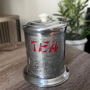 May include: A vintage chrome tea canister with a red interior. The word "TEA" is displayed in red block letters. The canister has a decorative embossed design and a white knob on the lid.