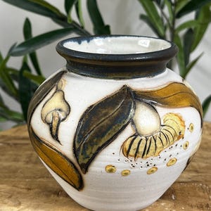 May include: A small, handcrafted ceramic vase with a white base and a dark blue rim. The vase features a hand-painted design of leaves and floral elements in shades of brown, yellow, and green. The vase is sitting on a wooden surface.