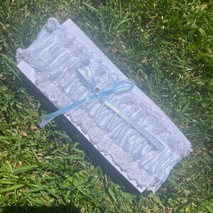 May include: A white satin garter featuring ruffled lace trim along the edges. The centre is adorned with a light blue ribbon tied into a bow, which includes a small, clear, circular rhinestone accent at the knot of the ribbon.