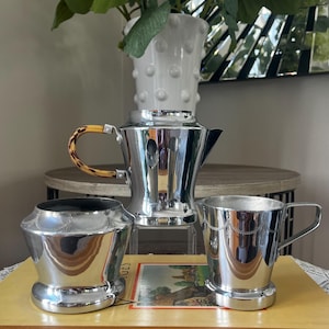 May include: A polished silver tea set, comprising a teapot with a bamboo-style handle, a sugar bowl, and a small cup. The set is displayed on a vintage book. The teapot is approximately 18 cm tall. A floral arrangement is in the background.