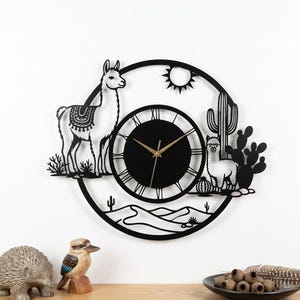 May include: Black metal wall clock featuring a desert scene with two llamas, cacti, and a sun. The clock has a round face with Roman numerals and gold-colored hands. The clock is mounted on a white wall.