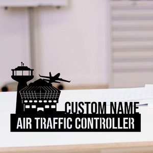 May include: Black metal desk sign featuring an airport control tower, a terminal, and a plane taking off. The sign includes the text "CUSTOM NAME" above "AIR TRAFFIC CONTROLLER". The sign is on a white desk.