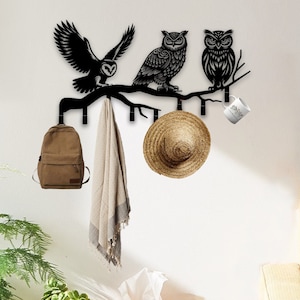 May include: Black metal owl wall hook with a branch design. The hook has five hooks and is holding a brown backpack, a beige scarf, a straw hat, and a white mug. The hook is mounted on a white wall.
